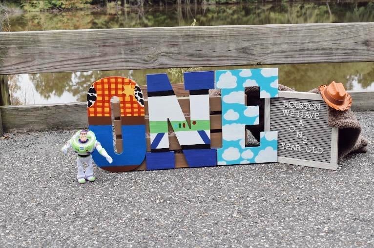 The word "One" is spelled with wooden letters, which are painted to match the clothes of Toy Story characters.